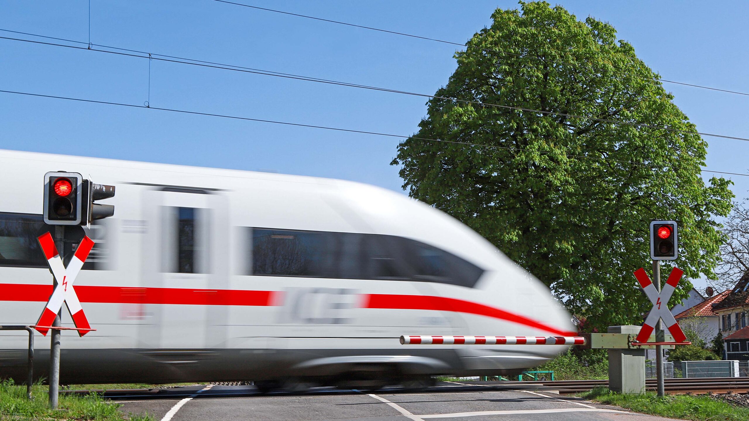 Realität statt Zukunft: Der Hochgeschwindigkeitszug ICE passiert einen beschrankten Bahnübergang an einer Landstraße. Moderne Infrastruktur mit Tunneln und Brücken machen den Schienenverkehr sicherer und beschleunigen ihn.